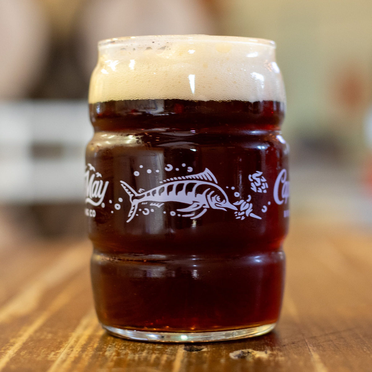 A Cape May Brewing can glass with a white marlin print, full of dark amber beer, on a wooden table