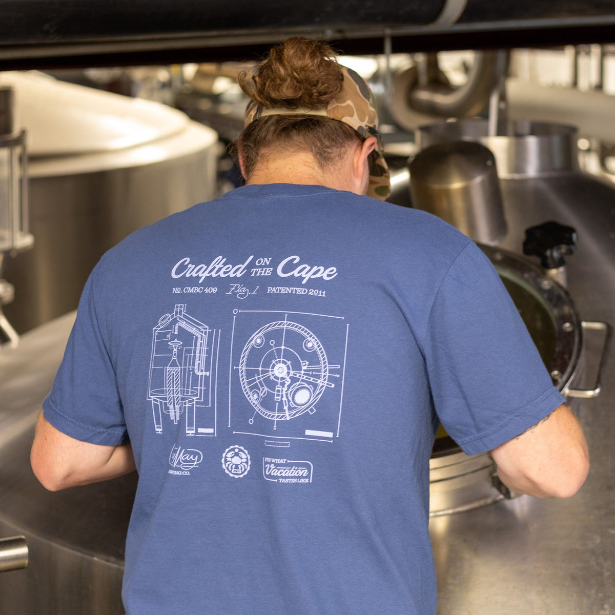 Back view of a person in a blue Cape May Brewing Crafted on the Cape tee, next to a steel brewing tank
