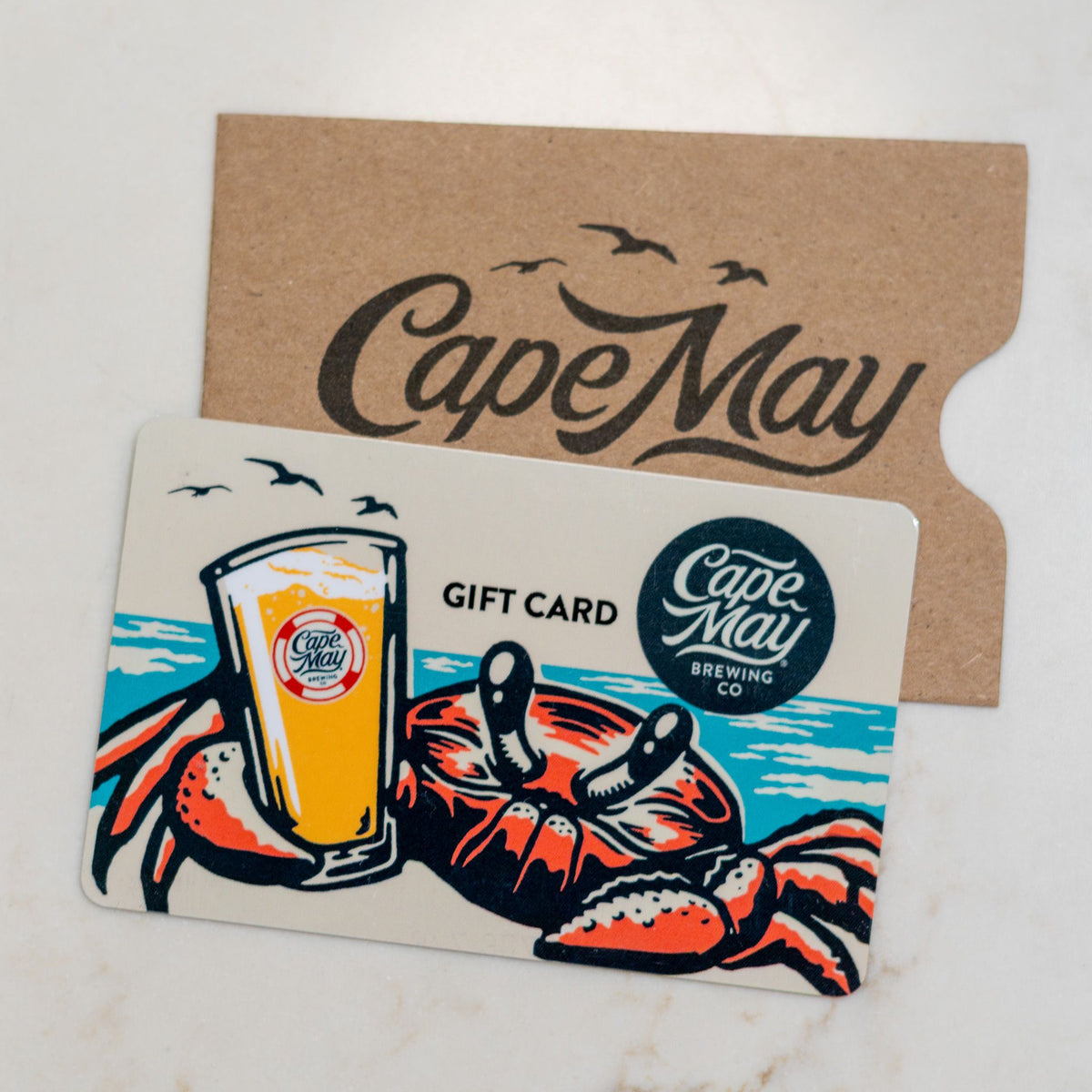 Cape May Brewing Co. gift card featuring a crab holding a pint of beer, resting on a kraft paper sleeve
